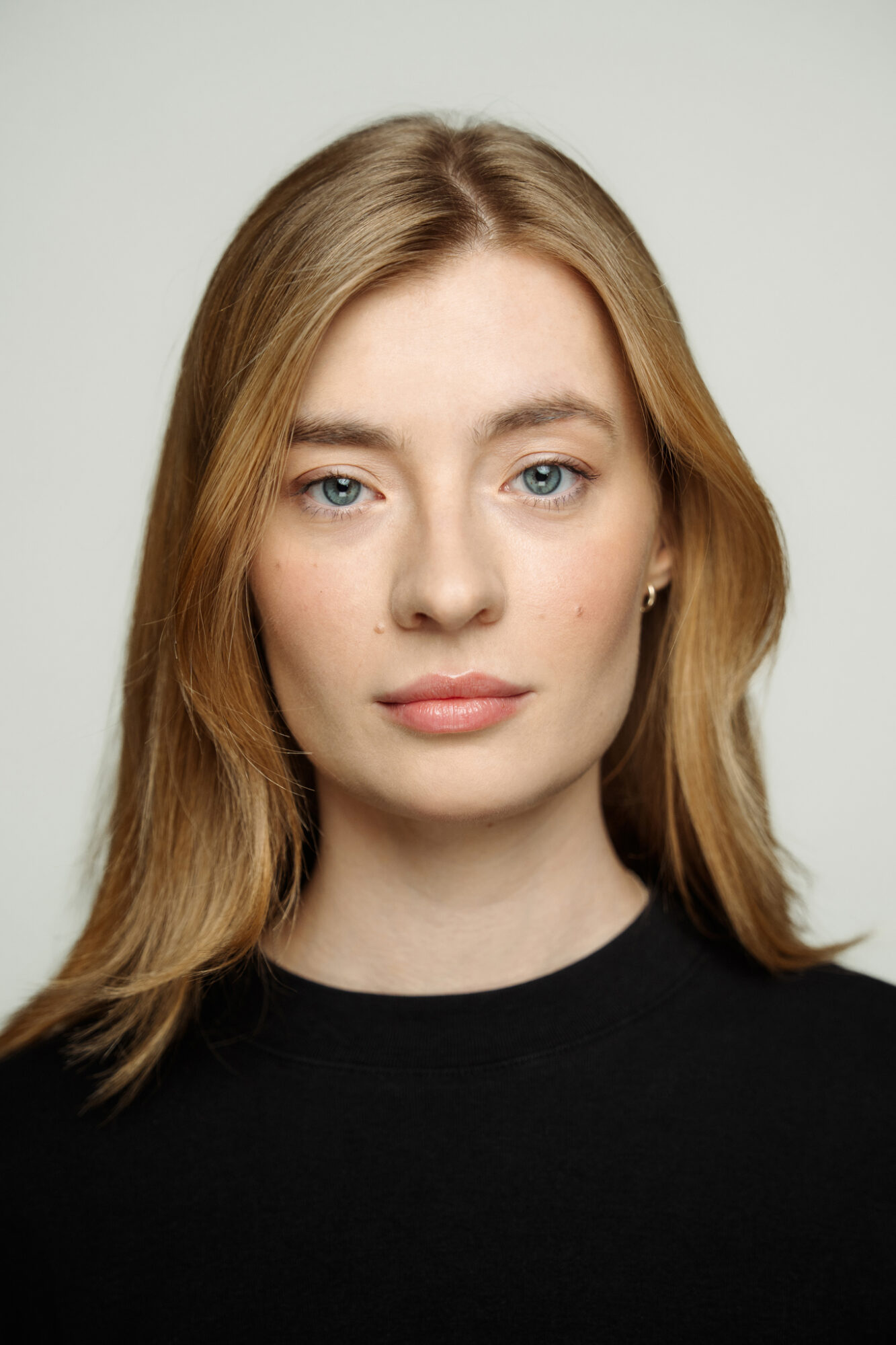 Portret niebieskookiej blondynki. Proste włosy spływają luźno za ramiona. Brwi gęste, policzki zaróżowione. 