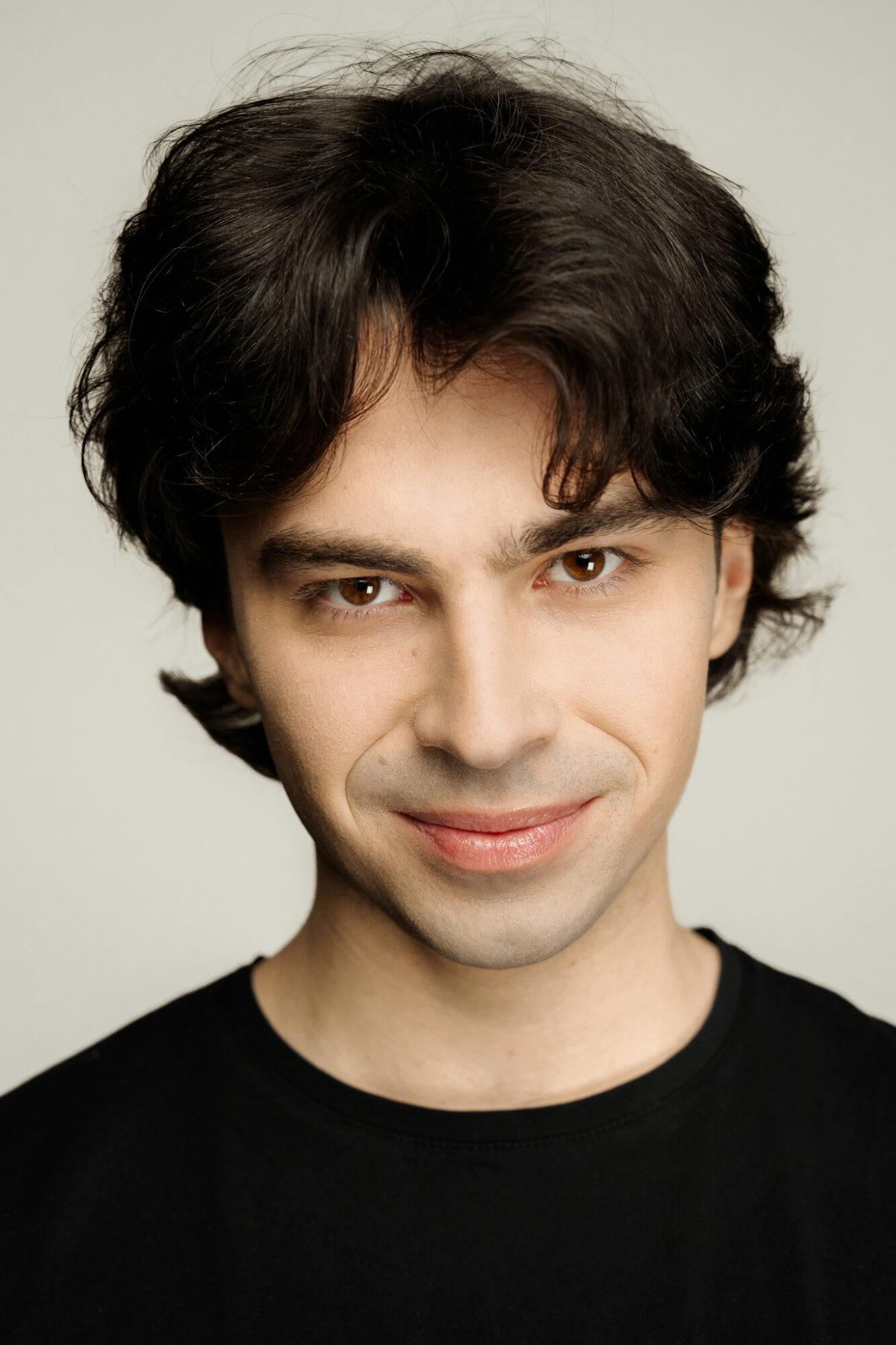 Portret mężczyzny. Ma ciemne, gęste włosy średniej długości, brązowe oczy. Lekko pochyla głowę do przodu, uśmiecha się.