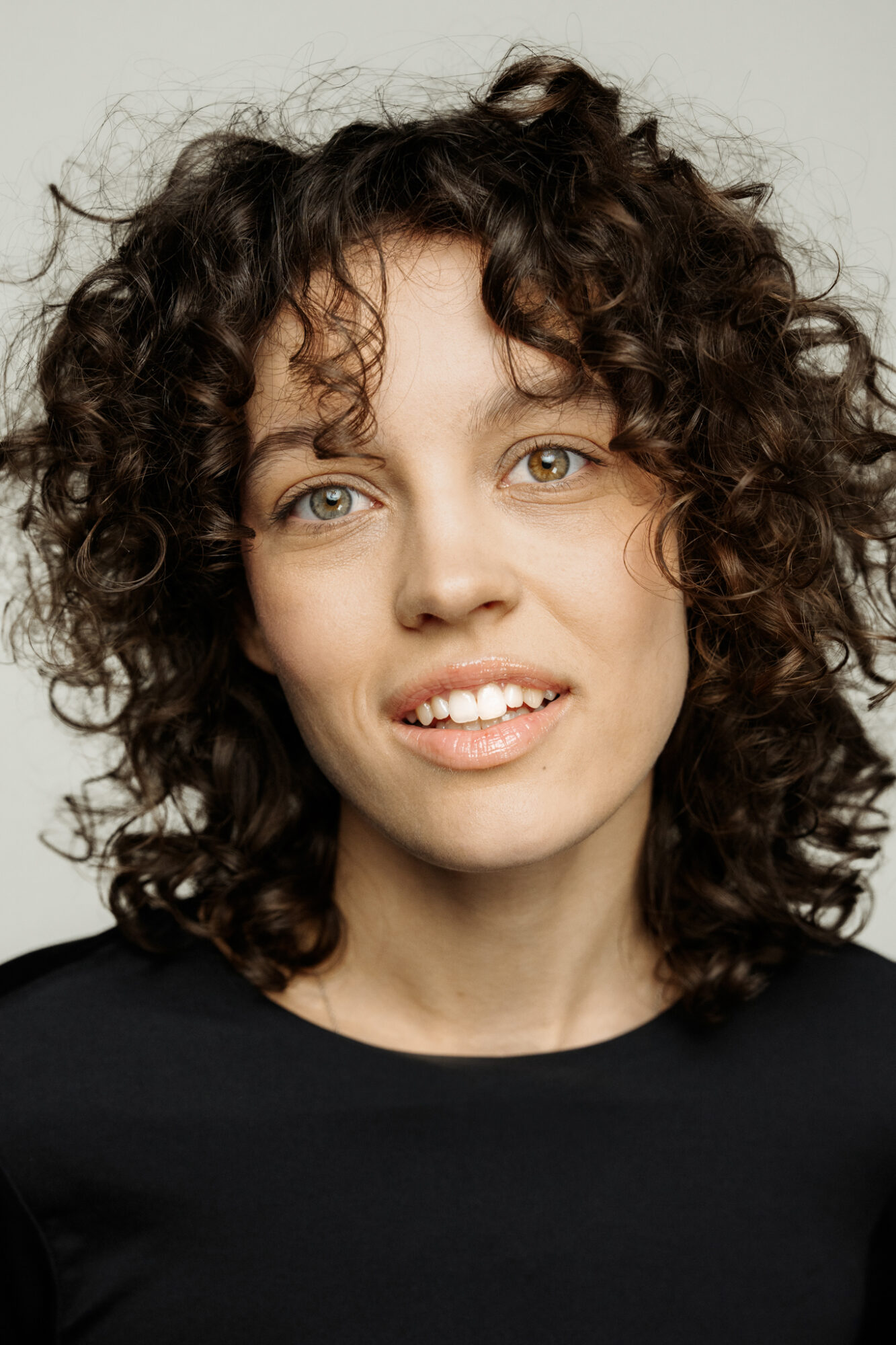 Portret szatynki z burzą loków. Ma szczupłą twarz, mocno zaznaczone kości policzkowe. Lekko przechyla głowę, rozchyla wargi w uśmiechu.