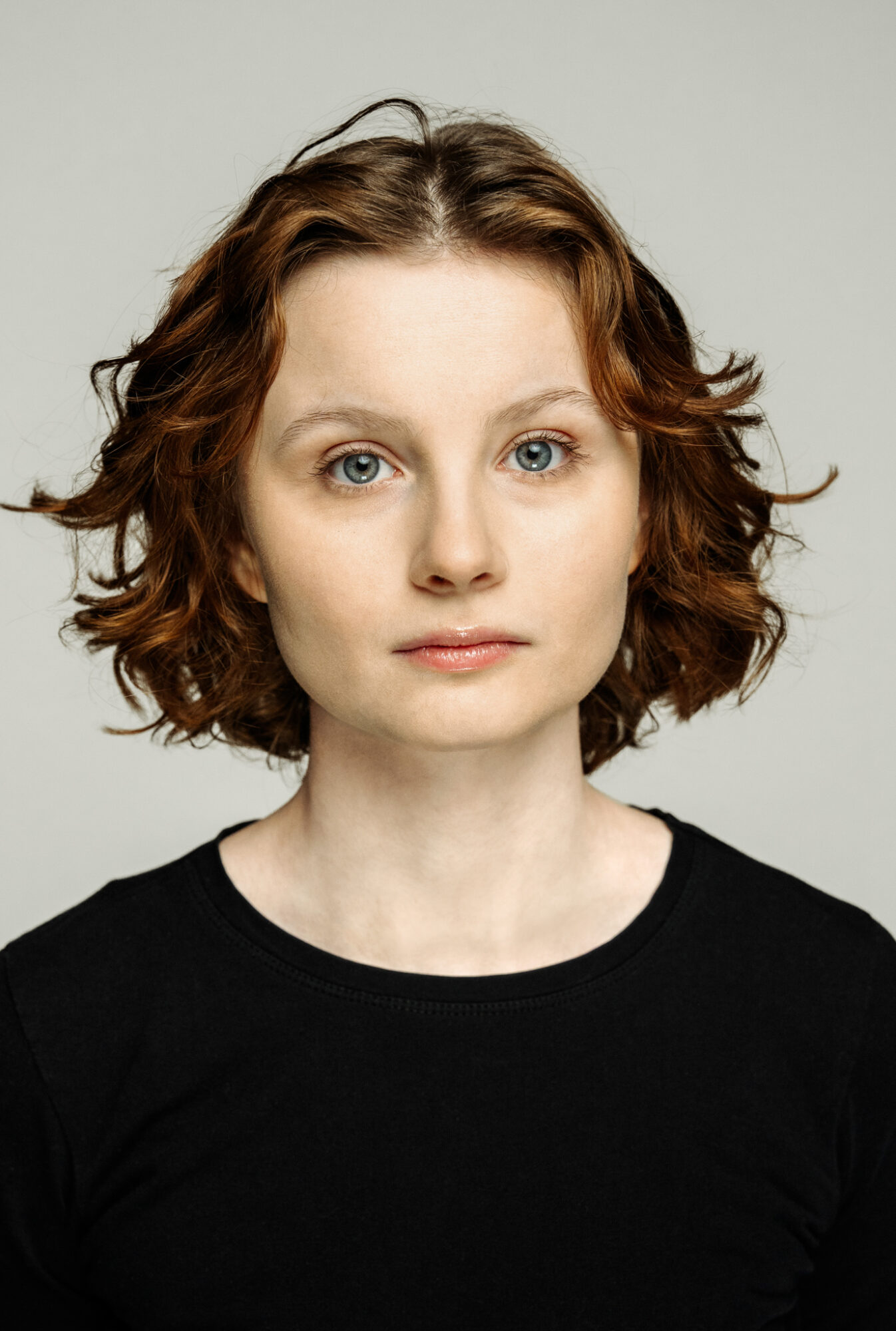 Portret młodej kobiety. Rude loki sięgają jej za ucho. Kobieta ma delikatne rysy, jasne brwi i duże, niebieskie oczy.