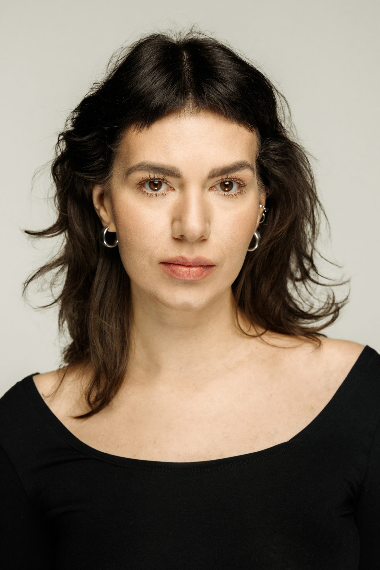 Portret ciemnowłosej kobiety. Ma brązowe oczy, gęste brwi. W uszach kilka srebrnych kolczyków.