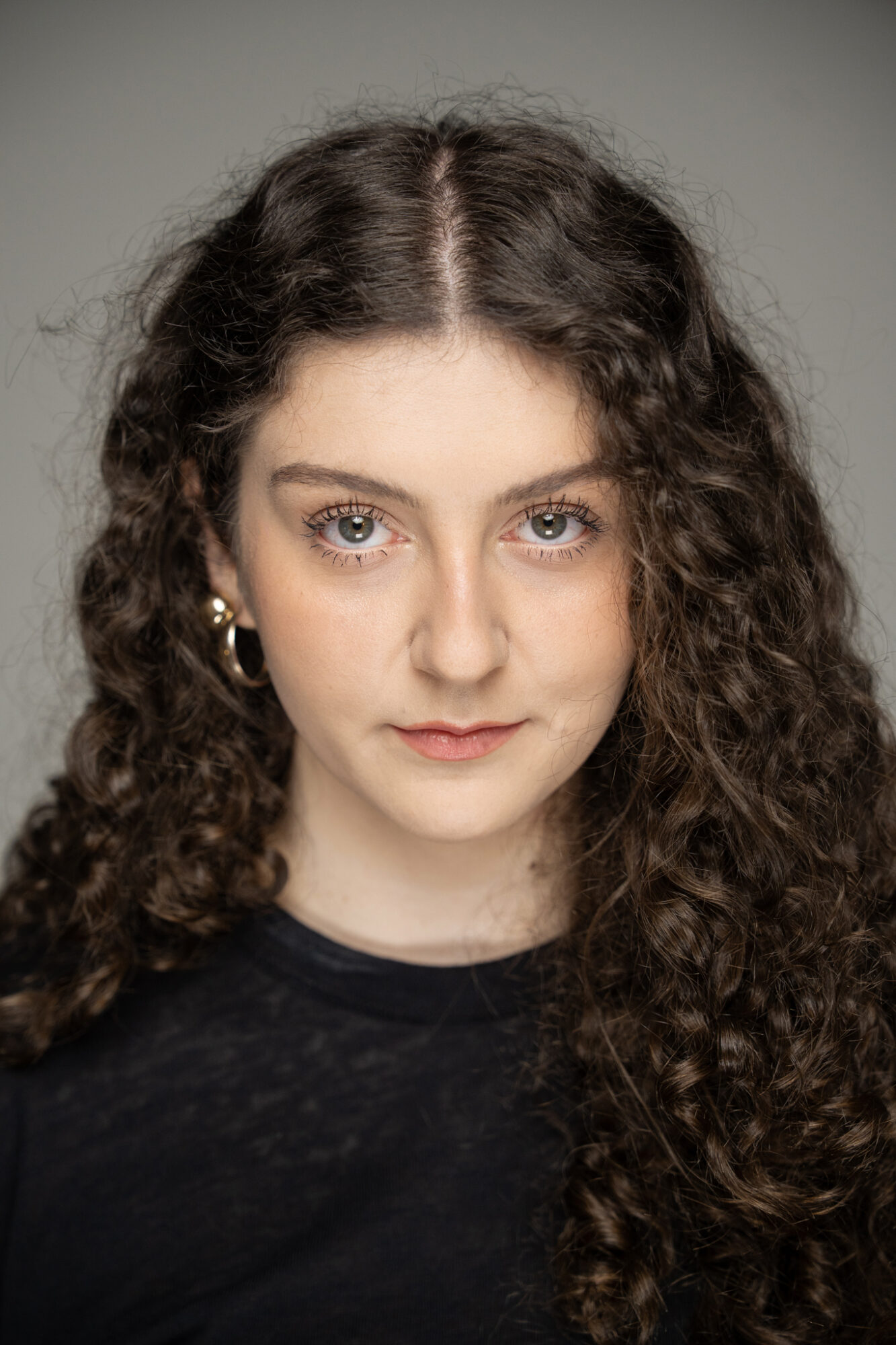 Portret kobiety z ciemnymi, kręconymi włosami. Lekko unosi kąciki ust.